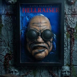 May include: A framed wall art piece featuring a sculpted, textured head with dark goggles and bared teeth, set against a blue background with the text CLIVE BARKER'S HELLRAISER. The frame is black, and the surrounding wall displays stone-like faces.