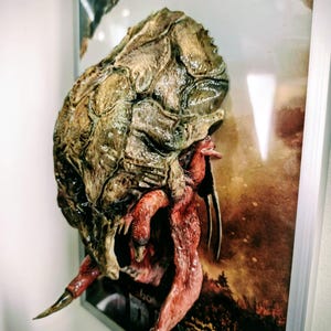 Könnte beinhalten: Detaillierte, dreidimensionale Skulptur eines Kreaturenkopfes mit einer strukturierten, braunen und grünen Panzerung und rotbraunen Anhängseln. Das Kunstwerk ist gerahmt, mit verschwommenem Hintergrund.