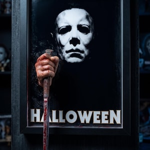 Könnte beinhalten: Ein gerahmtes 3D-Wandkunstwerk mit einer weißen Maske mit dunklen Augenhöhlen und einer Hand, die ein blutverschmiertes Messer hält und nach vorne ragt. Das Wort HALLOWEEN ist in weißen Großbuchstaben unten im schwarzen Rahmen gedruckt.