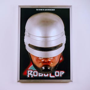 Könnte beinhalten: Gerahmtes Poster mit dem Gesicht von RoboCop. Das Poster zeigt einen silbernen Helm und ein schwarzes Visier. Das RoboCop-Logo ist in Rot und Blau gehalten. Der Text "THE FUTURE OF LAW ENFORCEMENT" steht oben.