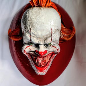 Op de afbeelding: Een wandsculptuur met het hoofd van een clown met een bleke witte huid, fel oranje haar en een brede, tandige grijns. De clown is bevestigd op een grote, glanzende, ballonvormige achtergrond met een dun rood touwtje dat onderaan hangt.