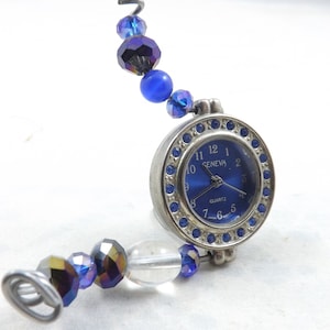 Puede incluir: Un reloj de cuarzo Geneva con esfera azul y bisel plateado, rodeado de cristales azules. El reloj está unido a una pulsera de cuentas con cuentas azules y transparentes.
