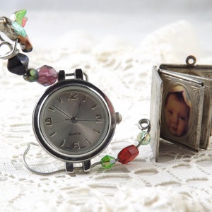 May include: A silver-toned quartz watch with a round face and a beaded hummingbird charm. A small, open, silver-toned locket with a baby's photo is next to the watch. The watch has a silver band and a beaded accent.