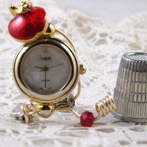 May include: A gold-toned quartz watch with a white face and the brand name "Staggle". The watch features a red apple-shaped embellishment and a red bead. A silver thimble is next to the watch.