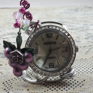 May include: Silver-toned Geneva Quartz watch with a mother-of-pearl face and crystal-studded bezel. The watch features a decorative grape cluster charm with purple and green accents, and a beaded accent. The watch is a unique jewelry piece.