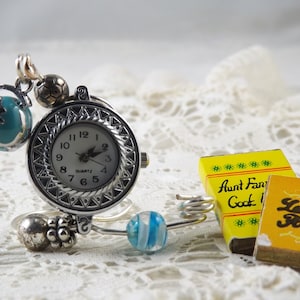 Puede incluir: Un reloj de pulsera plateado con esfera redonda y movimiento de cuarzo. El reloj presenta abalorios decorativos, como un globo terráqueo, un balón de fútbol y cuentas azules y blancas. También hay dos pequeños libros amarillos con texto.