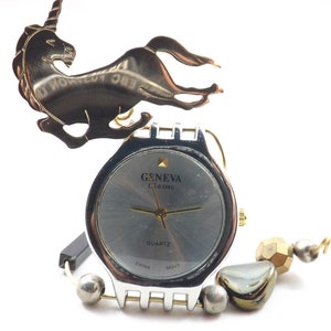 May include: A silver-toned Geneva Classic quartz watch with a gray face and a decorative unicorn charm. The watch has a silver band with decorative beads and a gold-toned spiral accent. The watch face reads "GENEVA Classic QUARTZ".