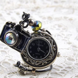 May include: A unique Geneva quartz watch with a black face and silver-tone filigree. A miniature black camera charm with a colorful lens is attached. The watch has a decorative bead and wire accents, creating a vintage, artistic aesthetic.
