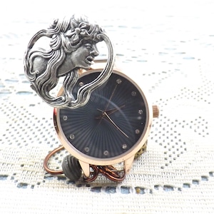 May include: A rose gold wristwatch with a dark blue face and silver accents. The watch features a decorative silver art nouveau style figure above the watch face. The watch has a copper-colored wire base.