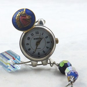 Puede incluir: Un reloj de cuarzo plateado con esfera blanca y la palabra "RUMOURS" impresa. Un charm de unicornio azul y dorado está unido a la esfera. El reloj tiene una pulsera de cuentas con cuentas coloridas y un charm de cubo de cristal.