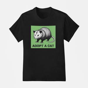 Pode incluir: Camiseta preta com um gr&aacute;fico de um gamb&aacute; em um quadrado verde, com o texto branco "ADOPT A CAT". O gamb&aacute; &eacute; retratado em tons de cinza e branco, com uma cauda longa. A camiseta tem gola redonda.