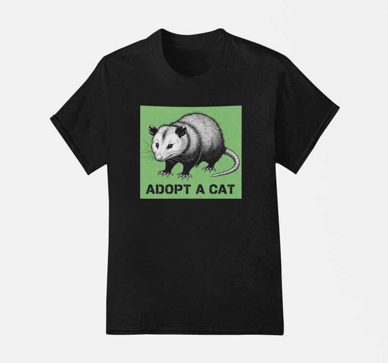 Pode incluir: Camiseta preta com um gr&aacute;fico de um gamb&aacute; em um quadrado verde, com o texto branco "ADOPT A CAT". O gamb&aacute; &eacute; retratado em tons de cinza e branco, com uma cauda longa. A camiseta tem gola redonda.