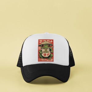 May include: A black and white trucker hat with a white front panel featuring a cartoon frog wearing a conical hat and eating ramen. The frog is green, and the ramen bowl is red. The hat has a curved brim.