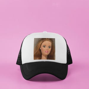May include: Black and white trucker hat with a white front panel featuring a doll's face with a surprised expression. The hat has a curved black brim and a mesh back, set against a pink background.