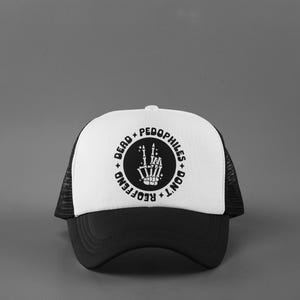May include: A black and white trucker hat with a white front panel and a black brim and mesh back. The front panel features a circular graphic with the text "DEAD PEDOPHILES DON'T REOFFEND" surrounding a skeleton hand making a peace sign.