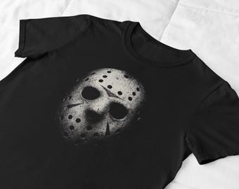 May include: Black t-shirt featuring a distressed graphic of a hockey mask with dark eye holes and numerous small, evenly spaced holes. The mask is a light gray color, and the shirt is made of a soft material.