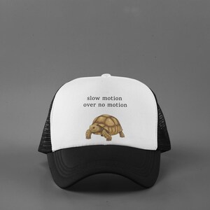 May include: A black and white trucker hat with a white front panel featuring the text "slow motion over no motion" above a tortoise illustration. The hat has a curved black brim and a mesh back.