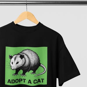 Pode incluir: Camiseta preta com uma ilustra&ccedil;&atilde;o em tons de cinza de um gamb&aacute; em um fundo verde. O texto "ADOPT A CAT" est&aacute; abaixo do gamb&aacute;. A camiseta est&aacute; pendurada em um cabide de madeira.