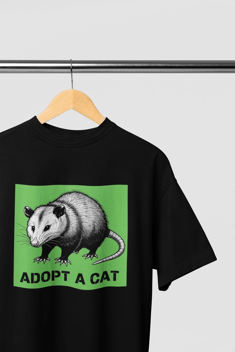 Pode incluir: Camiseta preta com uma ilustra&ccedil;&atilde;o em tons de cinza de um gamb&aacute; em um fundo verde. O texto "ADOPT A CAT" est&aacute; abaixo do gamb&aacute;. A camiseta est&aacute; pendurada em um cabide de madeira.