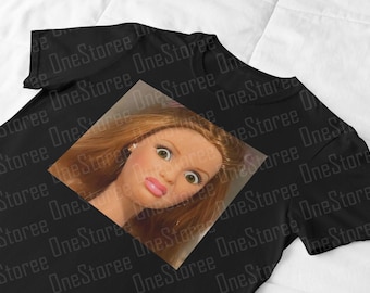 May include: Black t-shirt featuring a square image of a doll's face with wide eyes and pink lips. The doll has light brown hair and is wearing earrings. The shirt has a crew neck and short sleeves.