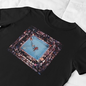 May include: Black t-shirt featuring a detailed image of a boxing ring from an aerial perspective. The image shows two boxers in the ring, surrounded by a crowd of spectators. The ring is blue, and the crowd is a mix of colors.