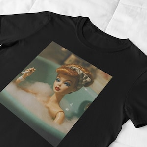 May include: Black t-shirt featuring a square image of a doll in a bathtub filled with bubbles. The doll has red hair, blue eyes, and red lipstick. The doll is holding a cigarette. The background is a light blue.