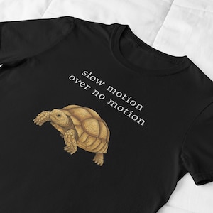 May include: Black t-shirt featuring a detailed illustration of a tortoise in shades of brown and tan. The text "slow motion over no motion" is printed in white. The shirt is made of a soft material.