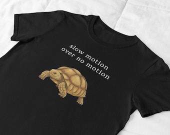 May include: Black t-shirt featuring a detailed illustration of a tortoise in shades of brown and tan. The text "slow motion over no motion" is printed in white. The shirt is made of a soft material.
