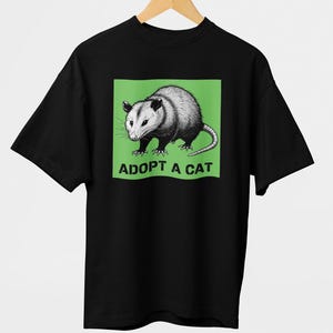 Pode incluir: T-shirt preto com uma ilustra&ccedil;&atilde;o detalhada de um gamb&aacute; dentro de um ret&acirc;ngulo verde. O texto "ADOPT A CAT" est&aacute; impresso abaixo do gamb&aacute;. O design est&aacute; centrado na camiseta.