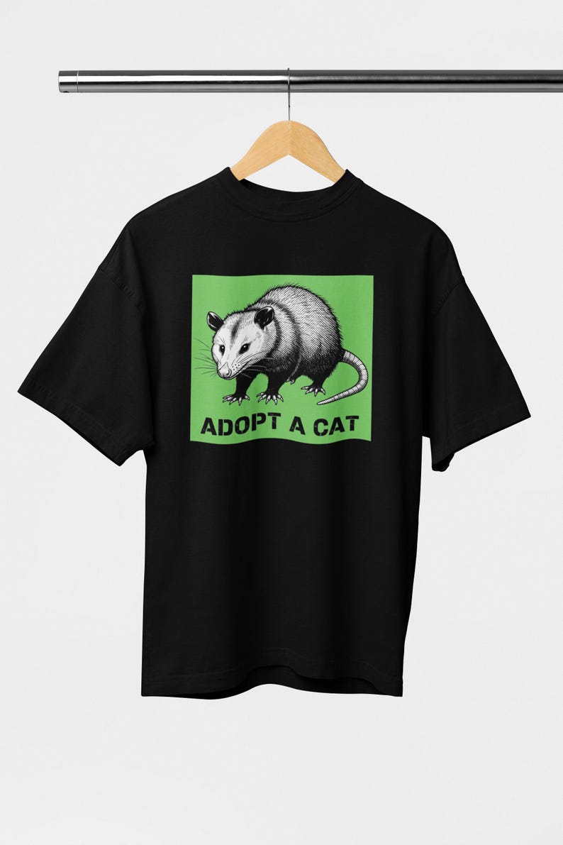 Pode incluir: T-shirt preto com uma ilustra&ccedil;&atilde;o detalhada de um gamb&aacute; dentro de um ret&acirc;ngulo verde. O texto "ADOPT A CAT" est&aacute; impresso abaixo do gamb&aacute;. O design est&aacute; centrado na camiseta.