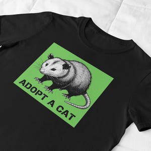 Pode incluir: Camiseta preta com um desenho de gamb&aacute; em um quadrado verde. O texto "ADOPT A CAT" est&aacute; impresso abaixo do gamb&aacute;. O design est&aacute; centralizado na camiseta.
