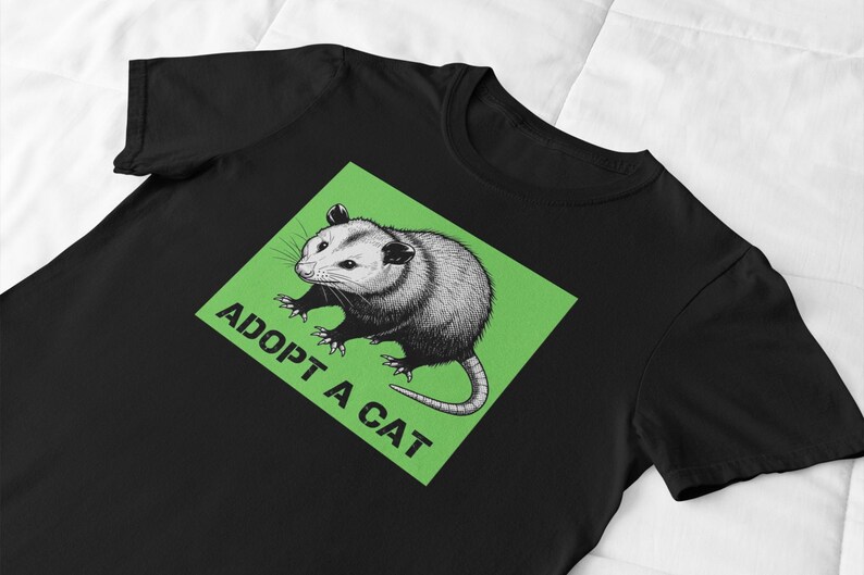 Pode incluir: Camiseta preta com um desenho de gamb&aacute; em um quadrado verde. O texto "ADOPT A CAT" est&aacute; impresso abaixo do gamb&aacute;. O design est&aacute; centralizado na camiseta.