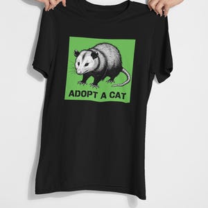 Pode incluir: Camiseta preta com uma ilustra&ccedil;&atilde;o de gamb&aacute; em um quadrado verde. O texto "ADOPT A CAT" est&aacute; impresso abaixo. A camiseta &eacute; segurada por duas m&atilde;os. O design &eacute; uma interpreta&ccedil;&atilde;o humor&iacute;stica da ado&ccedil;&atilde;o de animais.