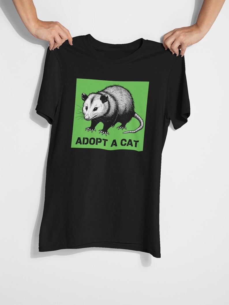 Pode incluir: Camiseta preta com uma ilustra&ccedil;&atilde;o de gamb&aacute; em um quadrado verde. O texto "ADOPT A CAT" est&aacute; impresso abaixo. A camiseta &eacute; segurada por duas m&atilde;os. O design &eacute; uma interpreta&ccedil;&atilde;o humor&iacute;stica da ado&ccedil;&atilde;o de animais.