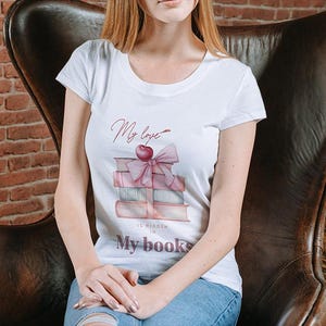 May include: White t-shirt with a round neck featuring a watercolor design of stacked books, a pink bow, and a cherry. The text "My love is hidden in My books" is printed on the shirt. The person is sitting in a brown leather chair.