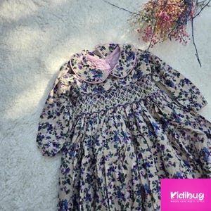 May include: A long-sleeved dress with a floral print in shades of purple and blue on a beige background. The dress has a smocked bodice and a pink Peter Pan collar.