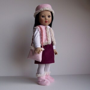 May include: A doll wearing a pink and white fur vest, a burgundy skirt, white tights, and pink boots. The doll is also wearing a pink hat with a white fur trim. The doll has brown hair and brown eyes.