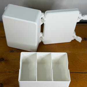 以下が含まれることがあります： 3つのコンパートメントとヒンジ付きの蓋が付いた白いプラスチック製の収納容器。蓋は開いており、内部が見えます。容器は木製の表面にあります。