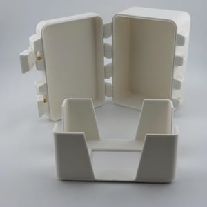 Könnte beinhalten: Eine weiße, rechteckige Box mit einem Klappdeckel, der geöffnet ist, um das Innere zu zeigen. Davor befindet sich ein separater, kleinerer weißer Behälter mit einer rechteckigen Öffnung. Box und Behälter bestehen aus glattem, mattem Material.