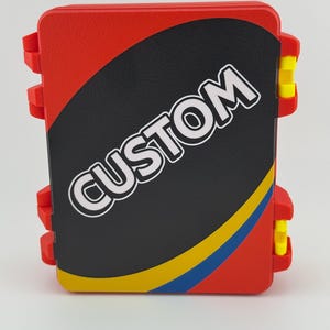 May include: A red rectangular case with the word "CUSTOM" in white lettering on a black oval. The case has yellow and blue stripes and yellow clasps. The case is designed to hold cards.