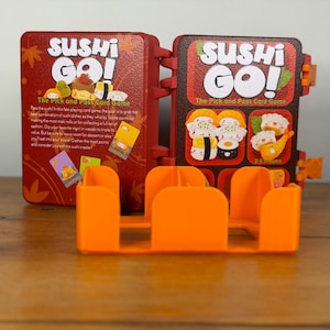 May include: A red and brown Sushi Go! card game box with orange card holders. The box features the game's title and sushi illustrations. The text "The Pick and Pass Card Game" is also visible.