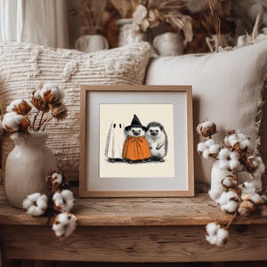May include: A framed print featuring a Halloween-themed illustration. The artwork depicts a ghost, a hedgehog in a witch costume, and another hedgehog. The print is in a wooden frame, resting on a wooden surface, with decorative cotton stems and pillows in the background.