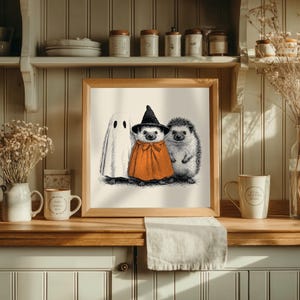 May include: A framed Halloween-themed illustration featuring a ghost, a hedgehog in an orange cape and witch hat, and another hedgehog. The artwork is in black and white with orange accents, perfect for seasonal decor.