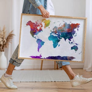 May include: A framed watercolour world map with vibrant colours. The map features continents in shades of red, yellow, blue, and purple. The wooden frame surrounds the map, which is held by a person.