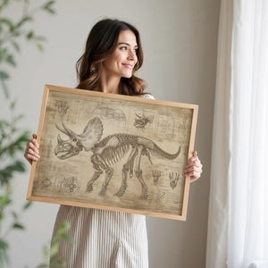 May include: A woman holds a framed print of a triceratops skeleton. The print is in a vintage style with a beige background and black lines. The triceratops skeleton is in the center of the print and is surrounded by diagrams and text.