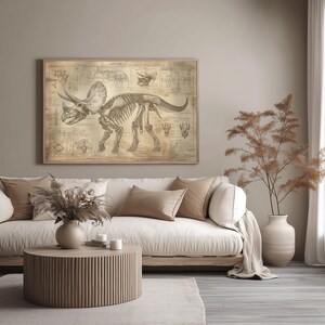 May include: A vintage-style illustration of a Triceratops skeleton on a beige background. The illustration is framed and hanging on a wall above a white couch with beige throw pillows. A potted plant with brown branches is on the right side of the image.
