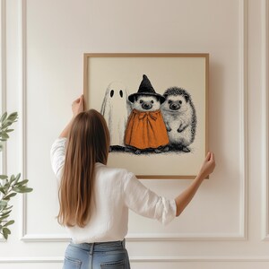 May include: A framed print featuring a Halloween theme. The artwork depicts a ghost, a hedgehog in a witch's hat and orange cape, and another hedgehog. The illustration is in black and white with orange accents, set against a cream background.