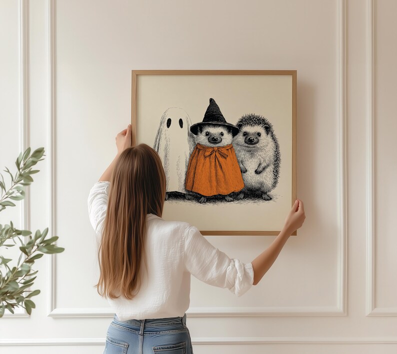 May include: A framed print featuring a Halloween theme. The artwork depicts a ghost, a hedgehog in a witch's hat and orange cape, and another hedgehog. The illustration is in black and white with orange accents, set against a cream background.