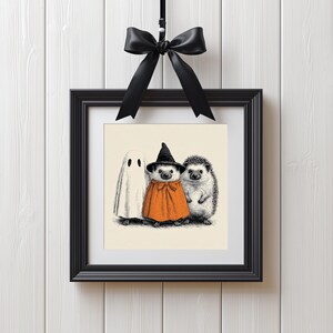 May include: A framed Halloween-themed print featuring a ghost and two hedgehogs. One hedgehog is dressed as a witch in an orange cape and black hat. The other hedgehog is in its natural state. The print is in a black frame and is suspended by a black ribbon.