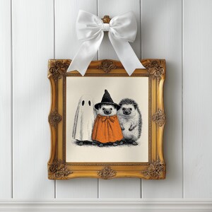 May include: A framed Halloween-themed print featuring a ghost, a hedgehog in an orange cape and witch hat, and another hedgehog. The ornate gold frame is topped with a white bow, creating a festive and whimsical display.